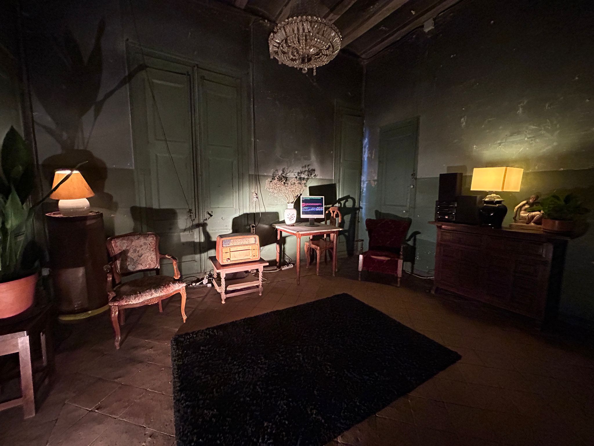 Photograph of the installation, a room full of old furniture including 2 old radios and a screen showing text